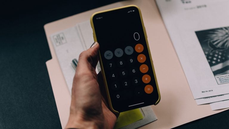 A hand holding a cell phone with the calculator app open. Underneath is a folder with financial documents on a table.